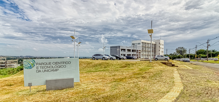 A foto mostra a placa do Parque Científico e Tecnológico da Unicamp e, ao fundo, prédios que fazem parte do Parque.