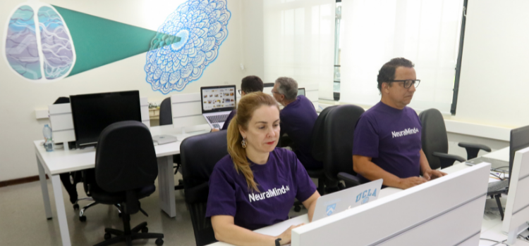 A foto é da sala de escritório da startup NerualMind, que fica no prédio Vértice do Parque Científico da Unicamp. Na foto há mesas com notebooks e pessoas trabalhando com a camiseta na cor roxa escrito NeuralMind. Na mesa da frente está Patrícia Tavares, CEO da Neuralmind.