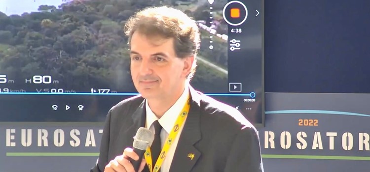 Foto de homem branco de cabelos encaracolados, veste terno preto e tem um cordão amarelo com um crachá branco ao redor do pescoço. Ele segura um microfone e está em frente a um painel com cores escuras onde aparece o nome do evento Eurosatory 2022. Fim da descrição.