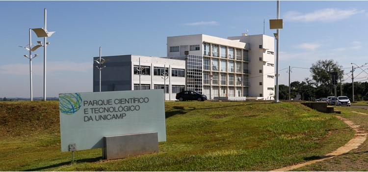 A imagem mostra a fachada do Parque Científico e Tecnológico da Unicamp. É possível ver uma placa com o nome do Parque e um de seus prédios. Fim da descrição.