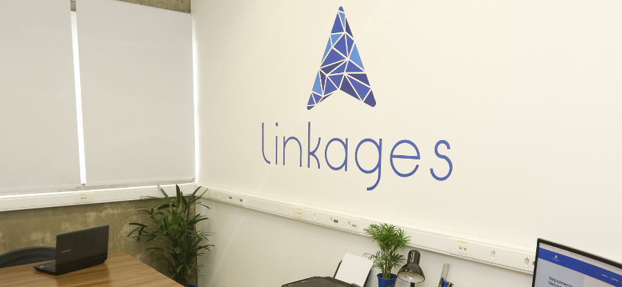 Imagem da sala de escritório da startup no Parque Científico da Unicamp. A imagem foca em uma parede branca com o logo da empresa, chamada Linkages. Fim da descrição.