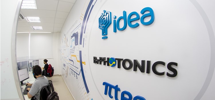 Foto de parede branca com desenhos que lembram um circuito de um chip em amarelo e azul e escrito o nome de três empresas que ocupam o prédio: Idea, BrPhotonics e Pi-Tec. Fim da descrição