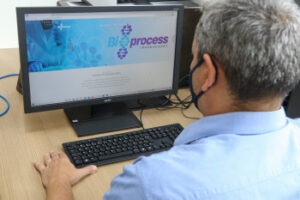 Homem branco, de cabelos grisalhos e camisa azul clara, mexe em computador de mesa que está logado na página da Bioprocess Improvement. Fim da descrição.