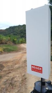 Foto em área externa mostra equipamento RuralMax, uma caixa retangular branca com o logotipo vermelho da Neger Telecom. Ao fundo uma estrada de terra e uma área arborizada. Fim da descrição.