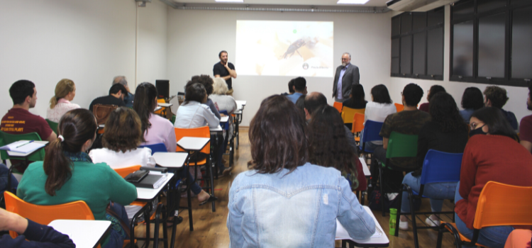 A imagem mostra uma sala de aula com pessoas sentadas em cadeiras em filas todas olhando para uma apresentação de slides com o palestrante à frente. Fim da descrição.