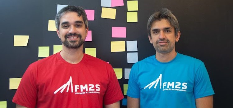 Murilo e Virgilio Marques dos Santos, fundadores da FM2S Educação e Consultoria, são homens brancos. Murilo usa barba e veste camiseta vermelha e Virgílio tem barba aparada e veste camiseta azul. Fim da descrição.