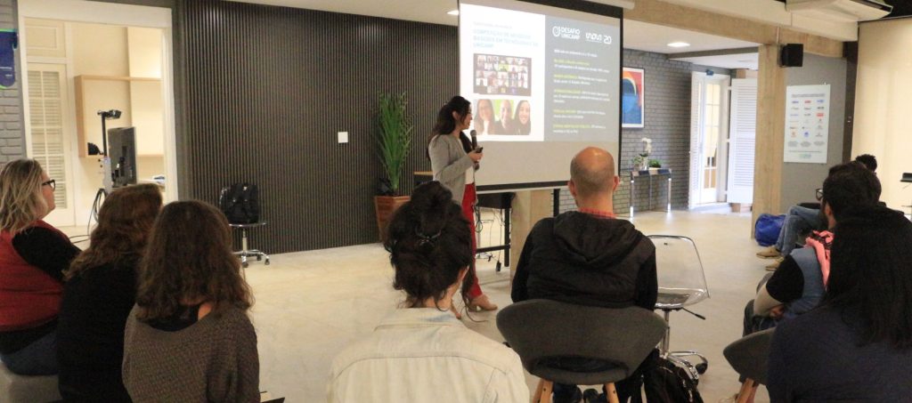 Foto de evento de apresentação da Inova. Mulher em pé com microfone apresenta um slide projeto e oito pessoas, de costas, sentadas, assistem. Fim da descrição.