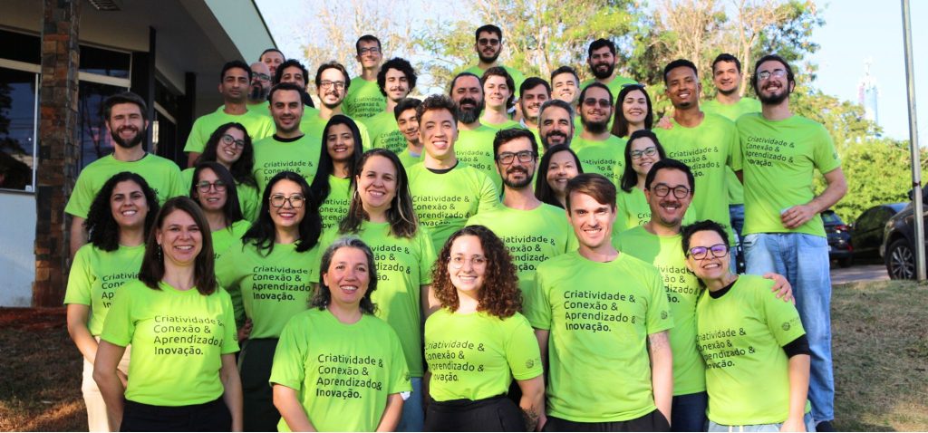 A imagem mostra o grupo de novos incubados posando para a foto. Todos estão utilizando uma camiseta verde com os seguintes dizeres: criatividade e conexão e aprendizado e inovação. Fim da descrição.