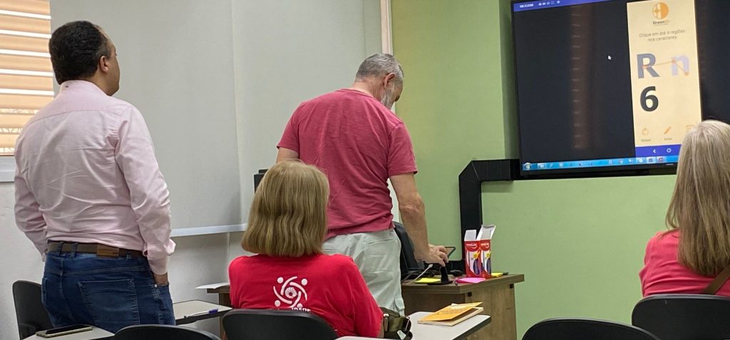 Professor vestido de camisa branca observando homem de camisa rosa testando o aplicativo no celular durante oficina realizada pela empresa-filha da Unicamp junto ao Programa UniversIDADE da Unicamp
