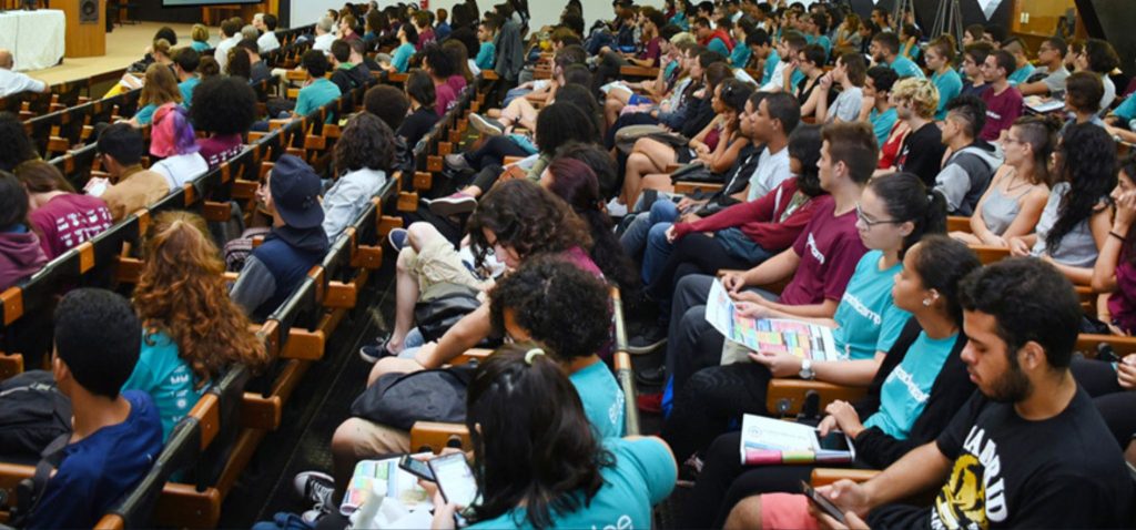 Foto colorida de centenas de alunos sentados em auditório da Unicamp, na recepção da calourada da Universidade. Fim da descrição.