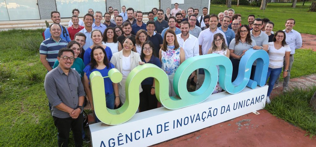 Foto colorida mostra dezenas de pessoas atrás do logo da Inova Unicamp. Fim da descrição.