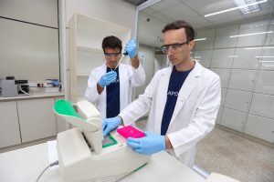 Fotografia. Lucas Fonseca e Lucas Parreiras estão em laboratório, usam jalecos brancos, luvas azuis e óculos de EPI, manuseiam instrumentos de laboratório. Fim da descrição.