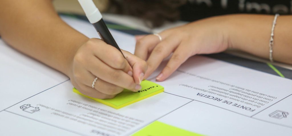 Foto colorida mostra braços de mulher branca escrevendo com canetão preto sobre papel colorido. Fim da descrição.