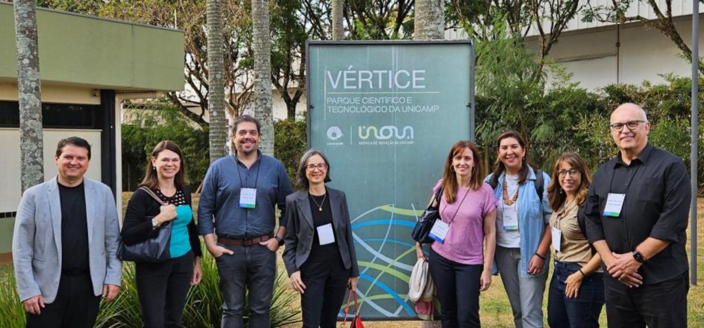 Foto posada dos participantes do congresso FGKD. Ao total são oito pessoas, em pé, olhando para a foto. Eles encontram-se em ambiente aberto, em frente à placa do Prédio Vértice, no Parque Científico e Tecnológico da Unicamp.