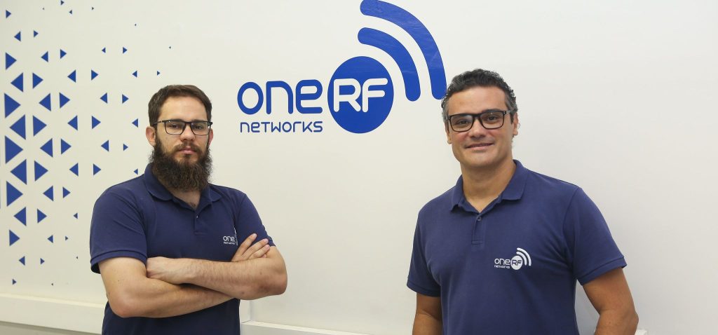 Foto posada de Gustavo Retuci e Eduardo Cardoso (esquerda para direita), fundadores da One RF. Eles encontram-se lado a lado, em frente ao logo da empresa. Eles vestem camiseta azul, com o logo da One RF. Fim da descrição.