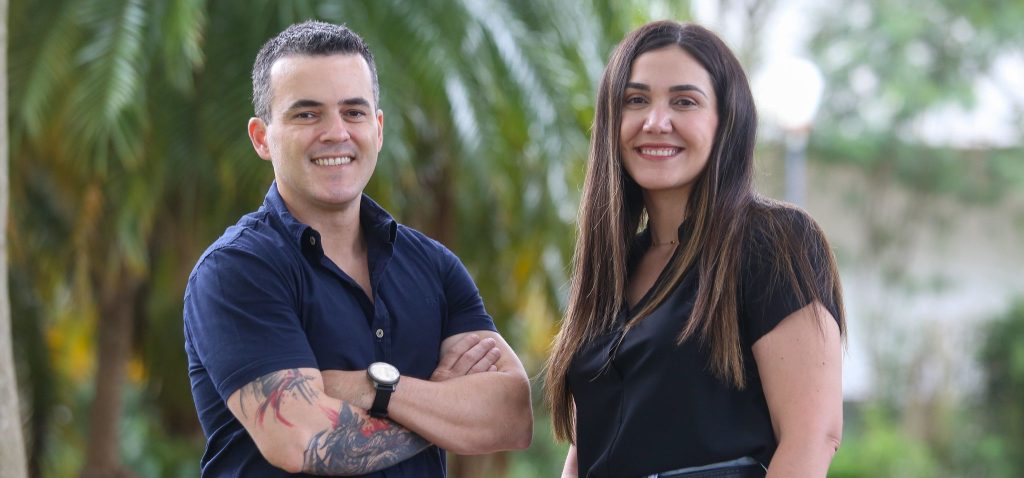 Foto posada dos fundadores da Inovia, Tiago Carvalho e Fernanda Balieiro. Na imagem, eles encontram-se olhando para a câmera, sorrindo, e contam com um árvores verdes ao fundo. O homem está de braços cruzados e veste uma camiseta azul marinho. A mulher, por sua vez, tem cabelos compridos e veste uma camiseta preta. Fim da descrição.