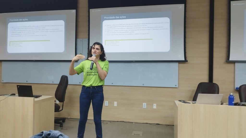 Anabelle Custódio em pé, vestindo camisa verde. Em segundo plano, apresentação sendo projetada Fim da descrição.