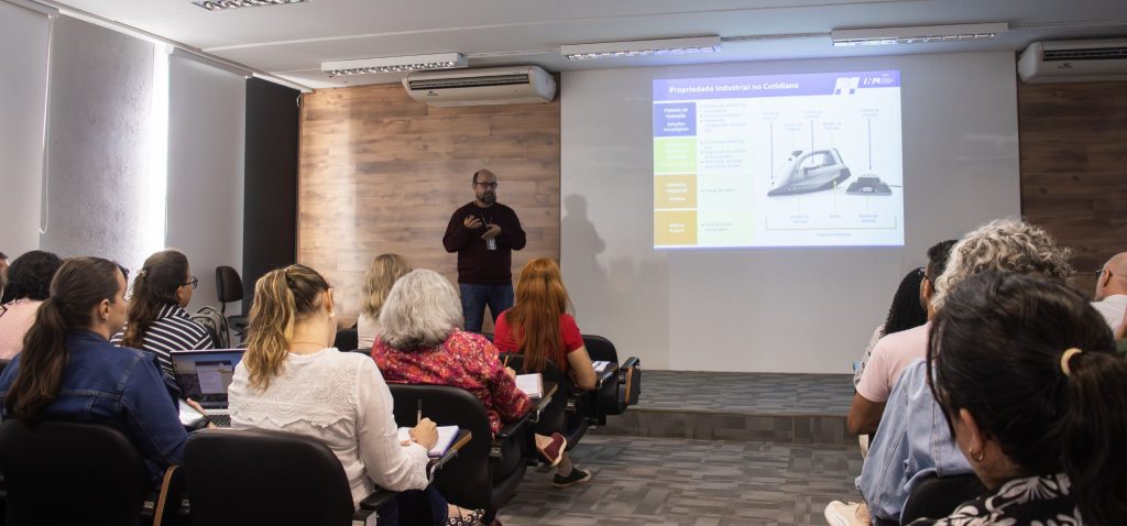 Foto feita em ambiente interno, no auditório da Incamp, traz o palestrante Clodoaldo Rulli ao fundo, ao lado de uma apresentação de slides projetada em tela. Em primeiro plano, está a plateia, de costas para o registro. Fim da descrição.