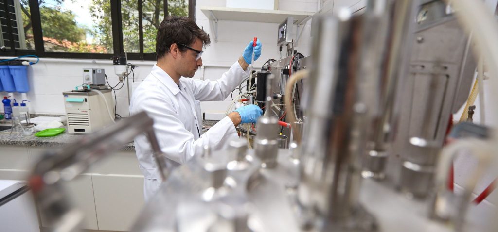 Foto interna de um homem manuseando instrumentos de laboratório. No registro, o homem encontra-se de lado para a câmera, em destaque, com elementos do laboratório em desfoque no primeiro plano. Ele veste jaleco branco, usa óculos e luvas azuis. Fim da descrição.