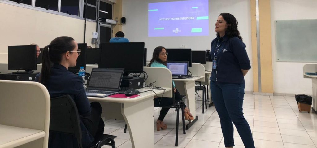 Sala de aula onde foi realizada a palestra. Duas mulheres sentadas e uma mulher em pé. Fim da descrição.