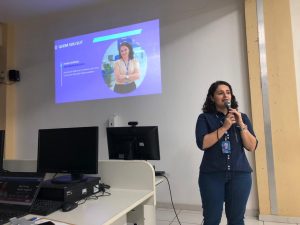 Em primeiro plano, Vivian Lourenço, analista de negócios do Sebrae. Ao fundo, projeção de sua apresentação. Fim da descrição.