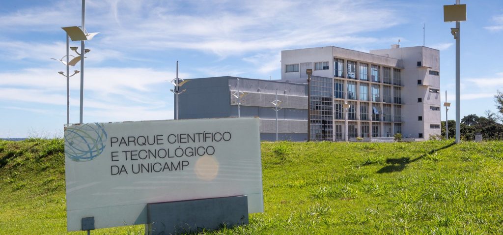 Foto externa do Parque Científico e Tecnológico da Unicamp. Imagem mostra o Prédio Núcleo, um dos edifícios que compõem a estrutura. No clique, feito em diagonal, é possível visualizar a placa indicando o nome do espaço. Há, ainda, um céu azul com nuvens brancas ao fundo e gramado verde. Fim da descrição.