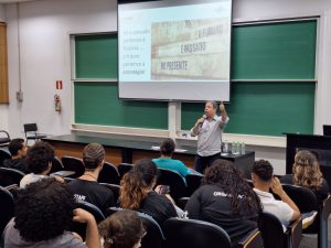 Anderson Santos, consultor do Sebrae, em segundo plano, está em pé palestrando à frente de sua apresentação. Em primeiro plano, de costas, estão alunos da Faculdade de Tecnologia da Unicamp. Fim da descrição. 