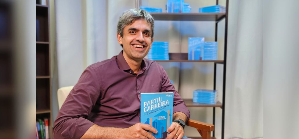 Foto posada de Virgílio Marques, autor do livro Partiu Carreira. Na imagem, ele encontra-se sentado, olhando para a câmera e segurando o livro lançado recentemente. Virgílio tem cabelo grisalho, veste uma camisa social e está sorrindo. Fim da descrição.