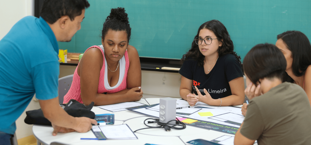 Na foto estão quatro mulheres sentadas envolta de uma mesa redonda prestando atenção na explicação do Vital, um homem, branco, com cabelos pretos. Ele está explicando sobre como montar um modelo de negócios usando o Canvas. A oficina aconteceu na Semana do Empreendedorismo de 2024. Fim da descrição.