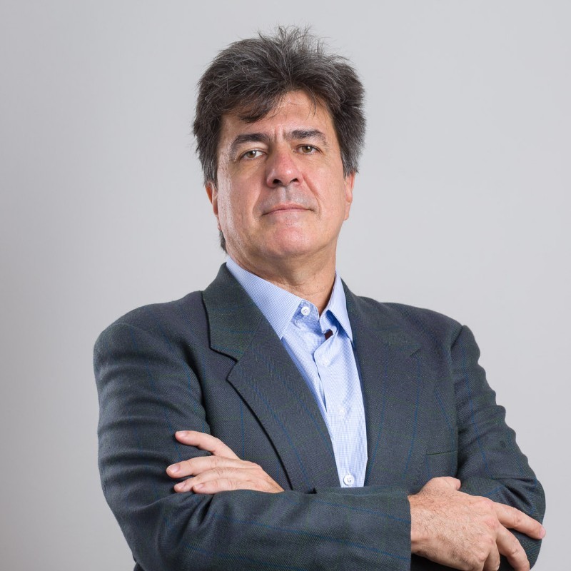 Retrato de meio corpo de um homem de cabelos escuros, vestindo um paletó cinza sobre uma camisa azul clara. Ele está com os braços cruzados e olha diretamente para a câmera contra um fundo cinza neutro. Fim da descrição.